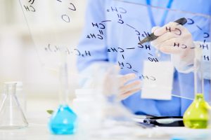 Unrecognizable chemist writing down structural formula on glass board while focused on work at modern laboratory, blurred background